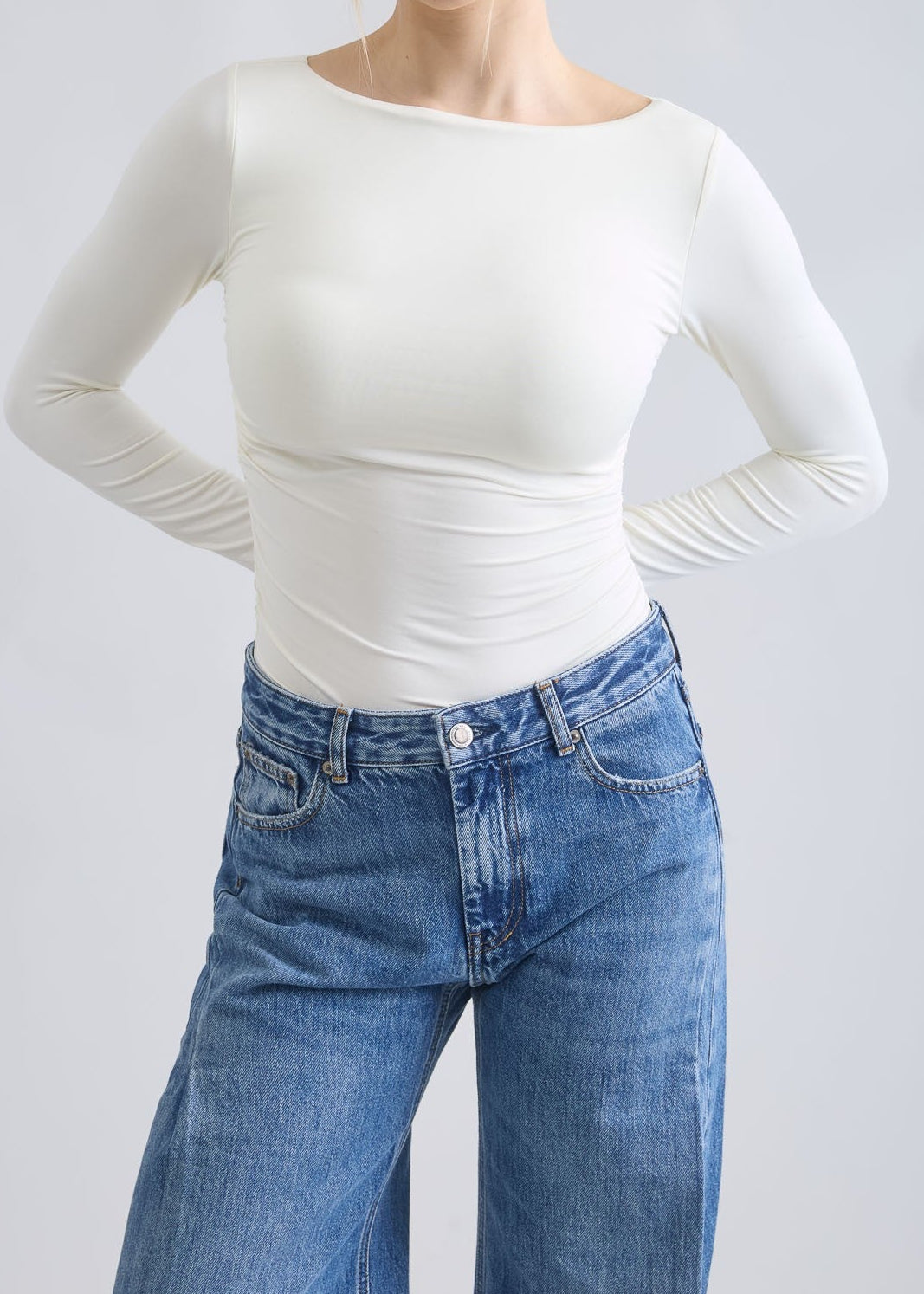 Woman wearing a white long-sleeve top and blue jeans against a plain background