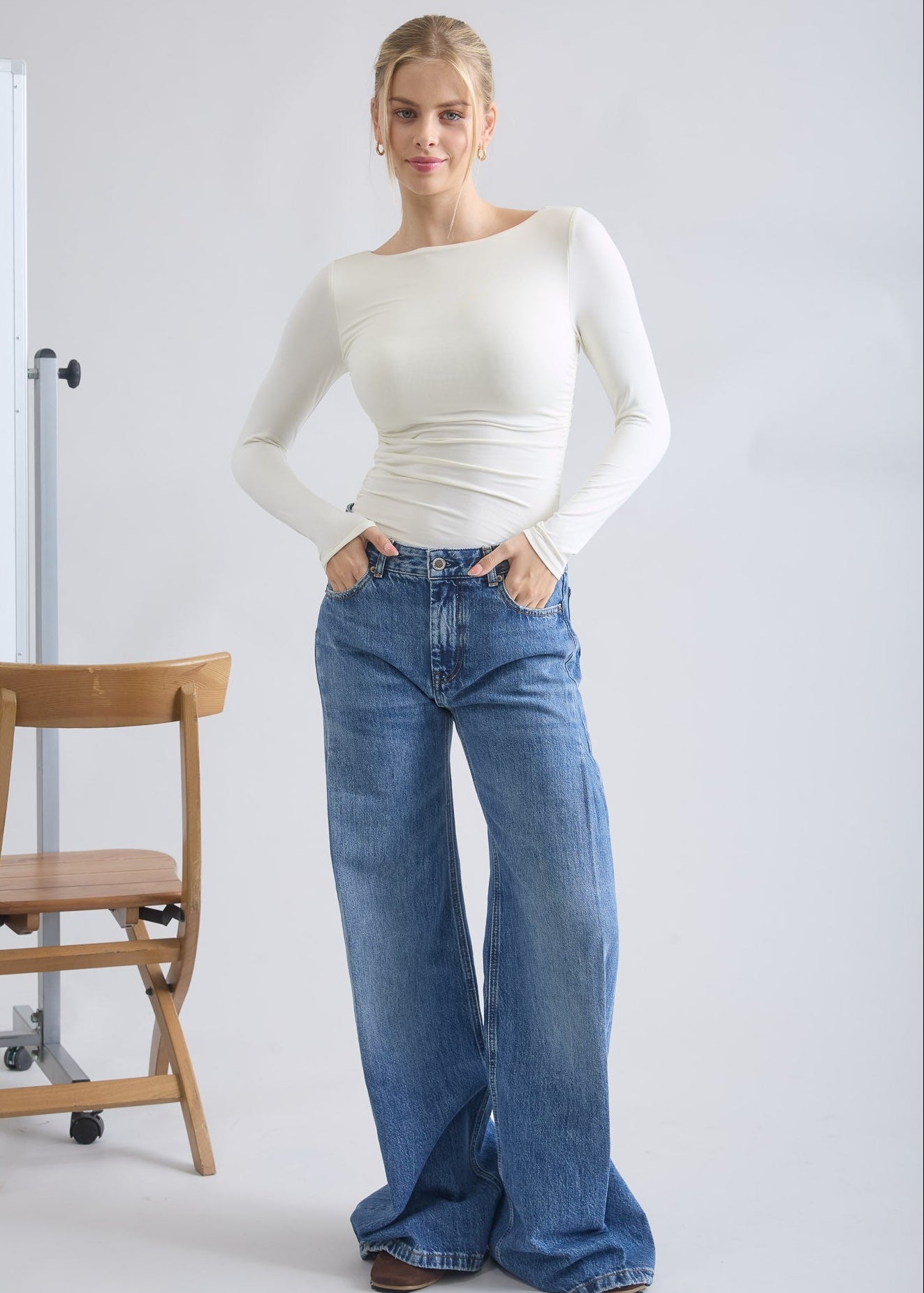 Woman wearing a white long-sleeve top and blue jeans standing against a plain background.