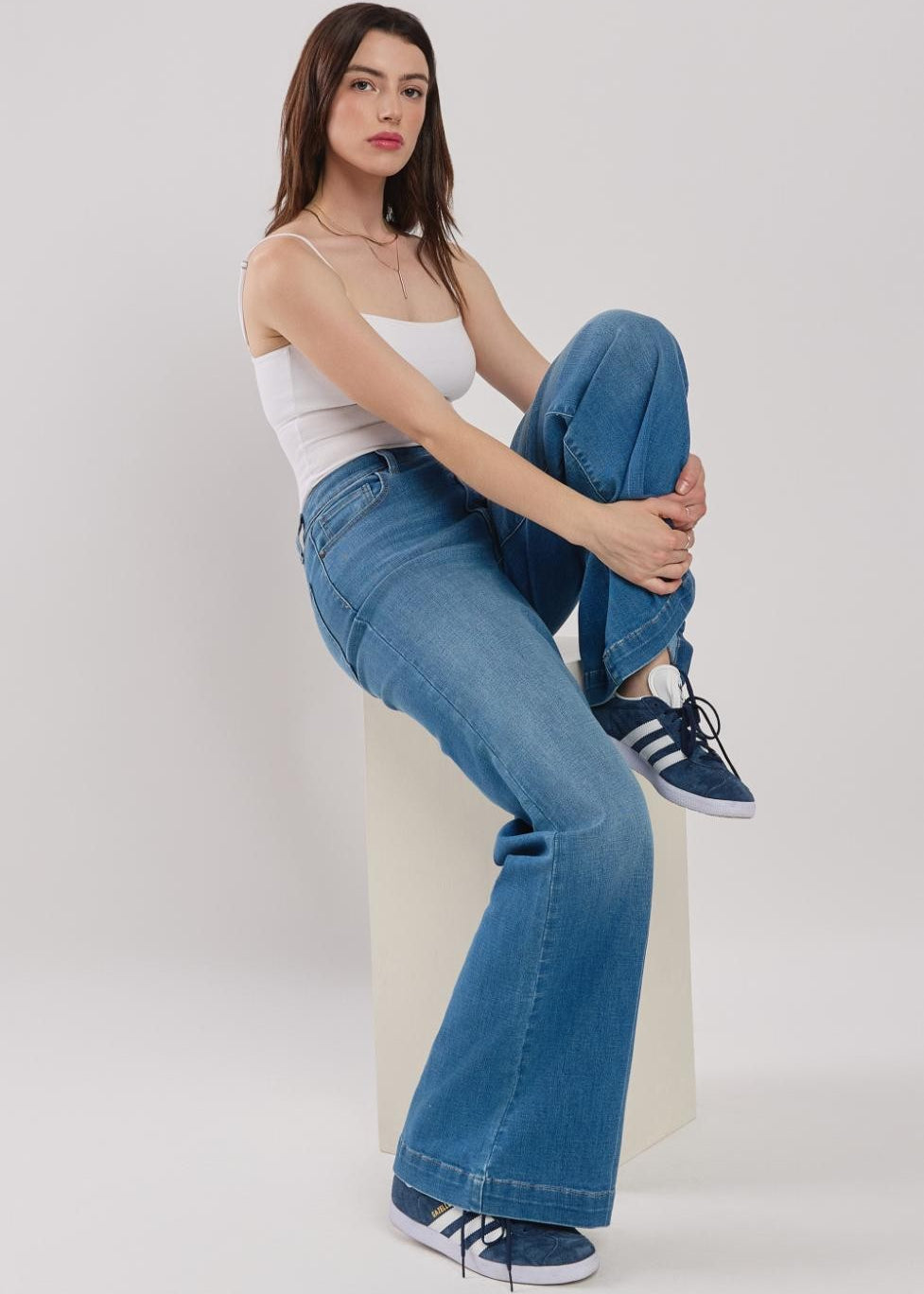 Woman sitting on a white stool wearing blue jeans and white sneakers against a plain background