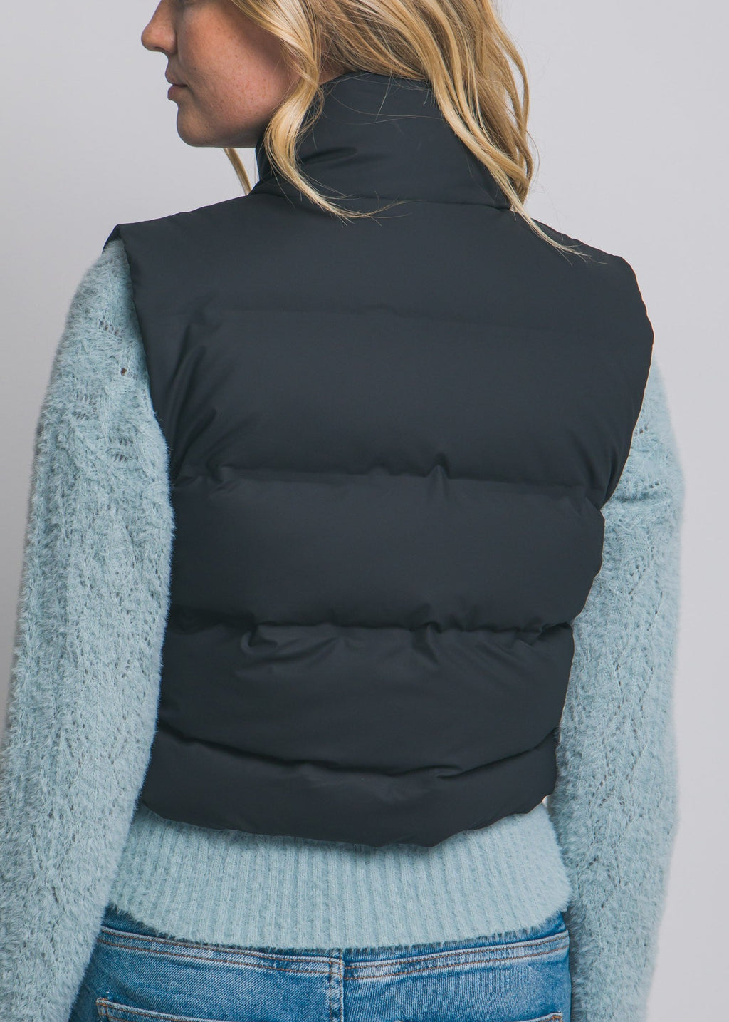 Person wearing a black puffer vest over a light blue sweater and jeans on a gray background