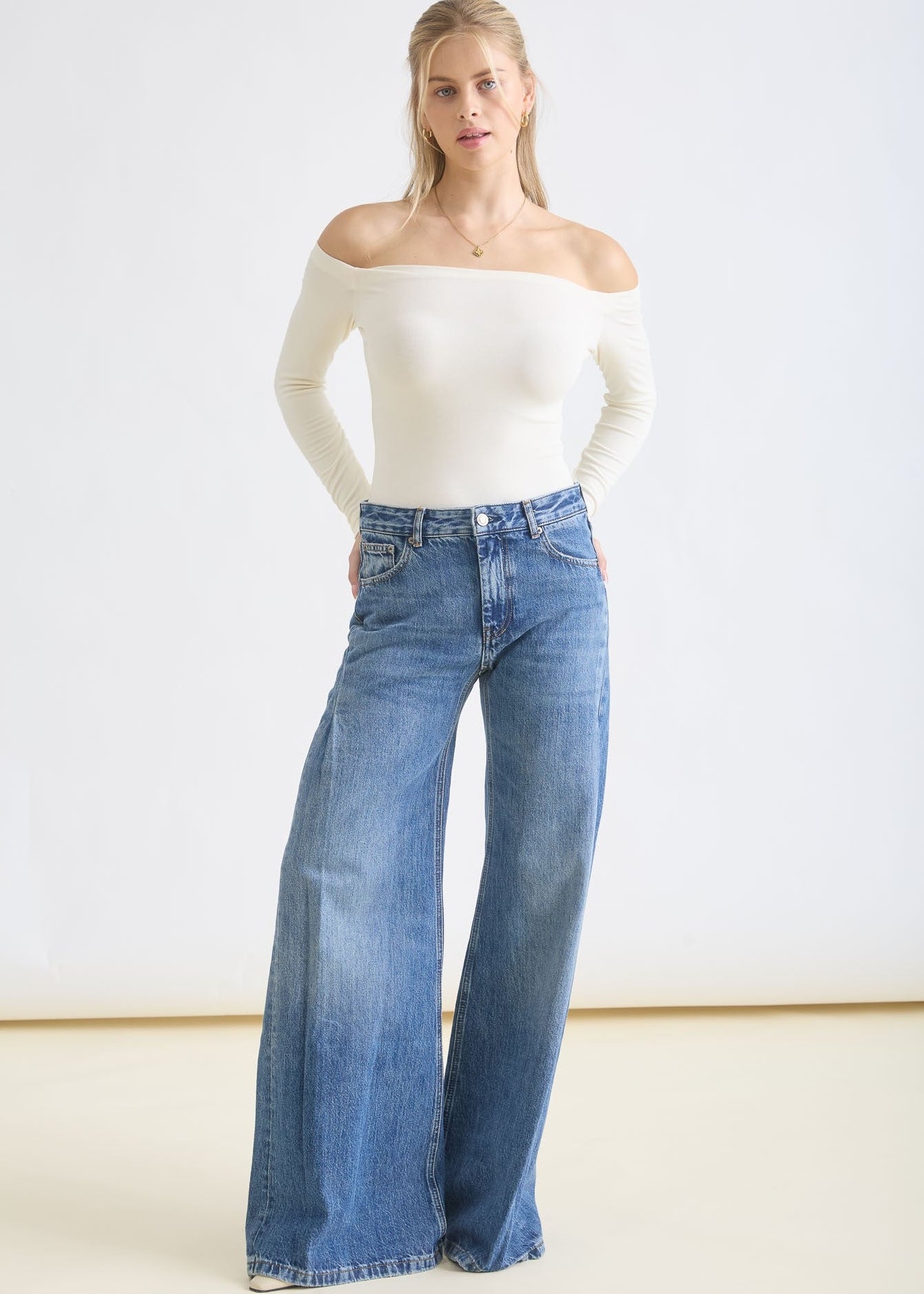 Woman wearing a white off-shoulder top and blue jeans on a plain background