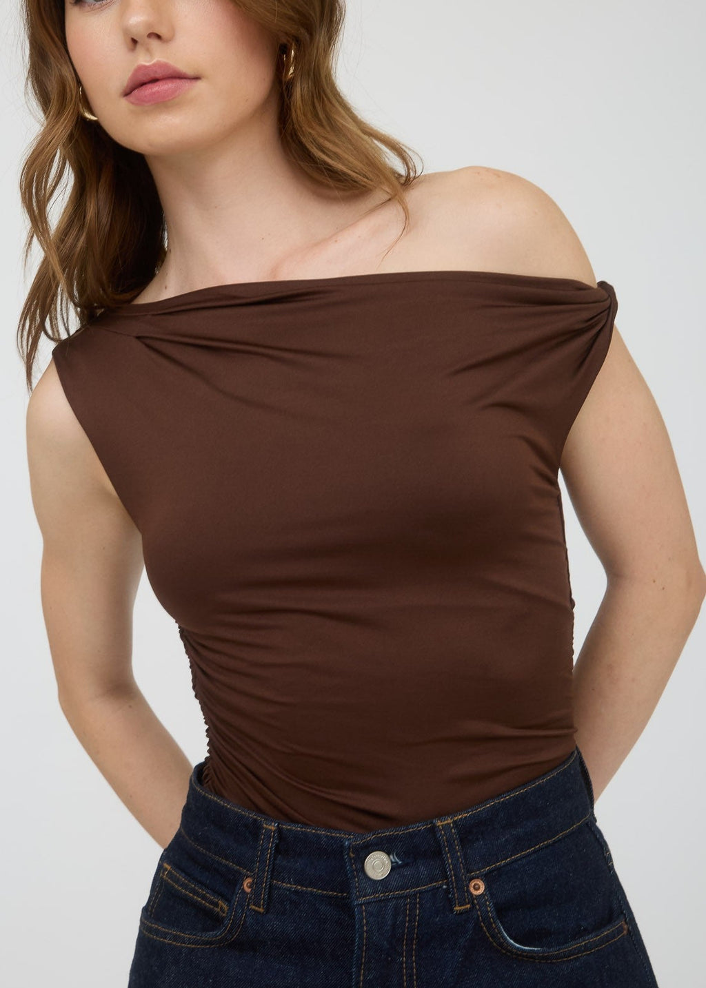 Woman wearing a brown off-shoulder top and dark jeans against a white background